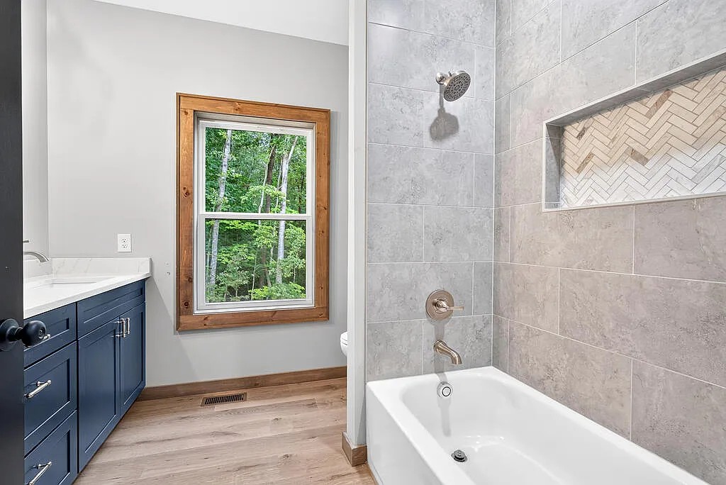 Bath with a navy vanity, wood-framed window, and tiled tub surround