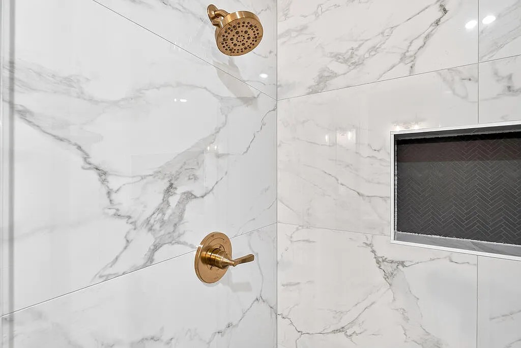 Walk-in shower with marble-look tile, gold fixtures, and a dark herringbone niche