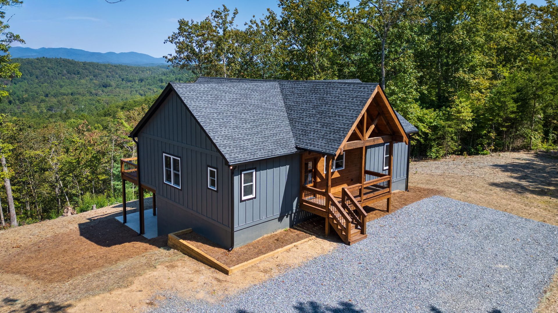 Side of a grey home with a timber-framed porch, gravel drive, and distant mountains