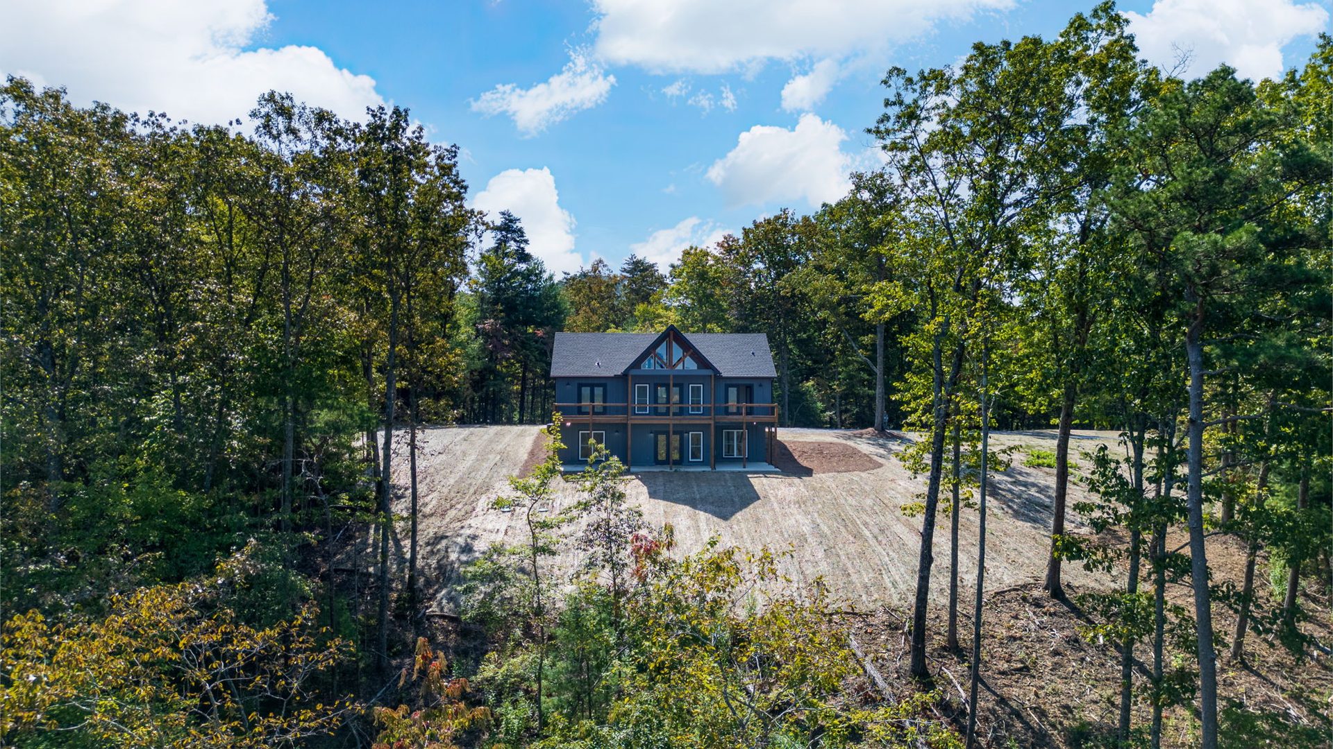 Aerial view of a grey home on a cleared gravel lot surrounded by dense forest