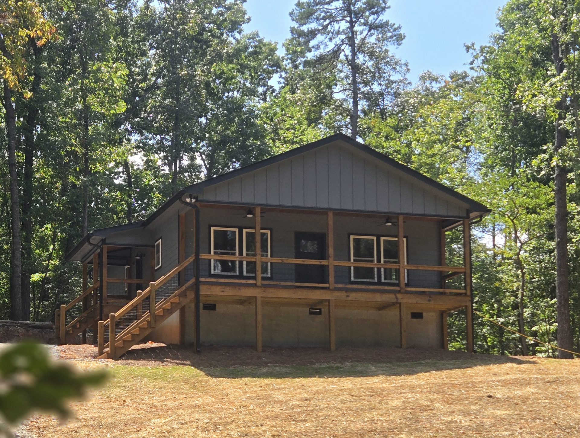 One-story gray-brown home with a full-length covered wood porch in a wooded setting