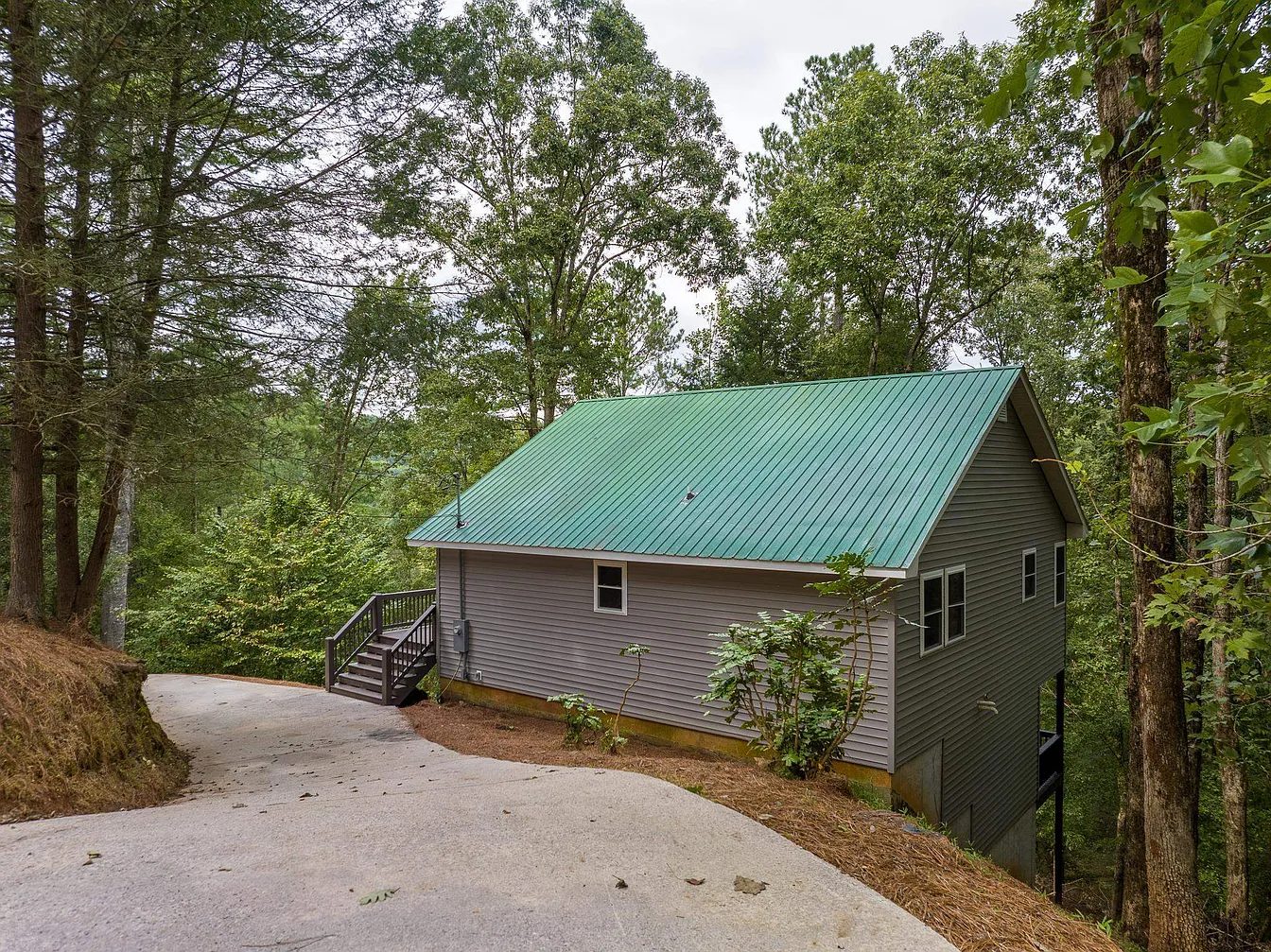 Two-story grey home with a green metal roof and wood side porch in the woods